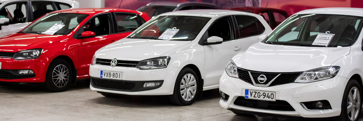 Menoauto Oy:n myyntihallissa esillä punainen ja kaksi valkoista autoa, joissa hintalaput tuulilaseissa.