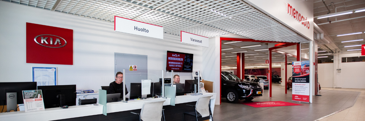 Menoauto Oy:n palvelutiski, jossa huolto- ja varaosapalvelut tarjolla. Taustalla näkyy autohalli ja myytäviä autoja.
