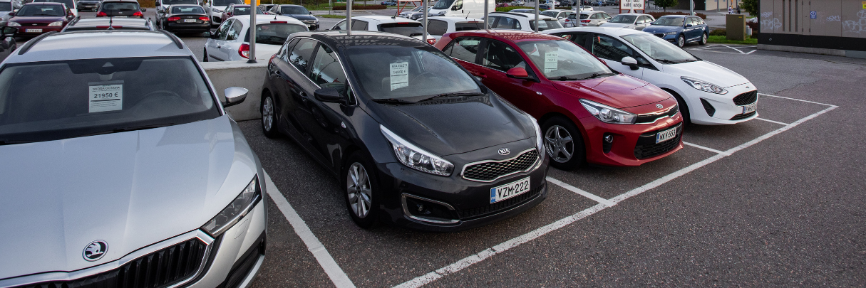 Useita myynnissä olevia autoja ulkona Menoauto Oy:n parkkipaikalla. Etualalla hopeinen, musta, punainen ja valkoinen henkilöauto, joissa hintatiedot esillä tuulilaseissa.