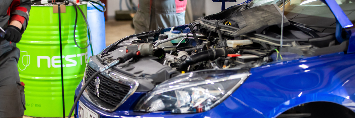 Menoauto Oy:n mekaanikko työskentelee sinisen auton parissa hallissa, konepelti avoinna ja huoltotyökaluja lähistöllä.