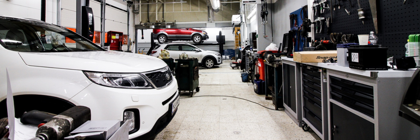 Tilava autokorjaamo, jossa lähellä kameraa on valkoinen SUV, punainen auto on hydraulisella nosturilla ja hopeanvärinen auto lattialla. Seinustoilla on työkaluja ja koneita korostamassa järjestelmällistä ja toimivaa työtilaa.